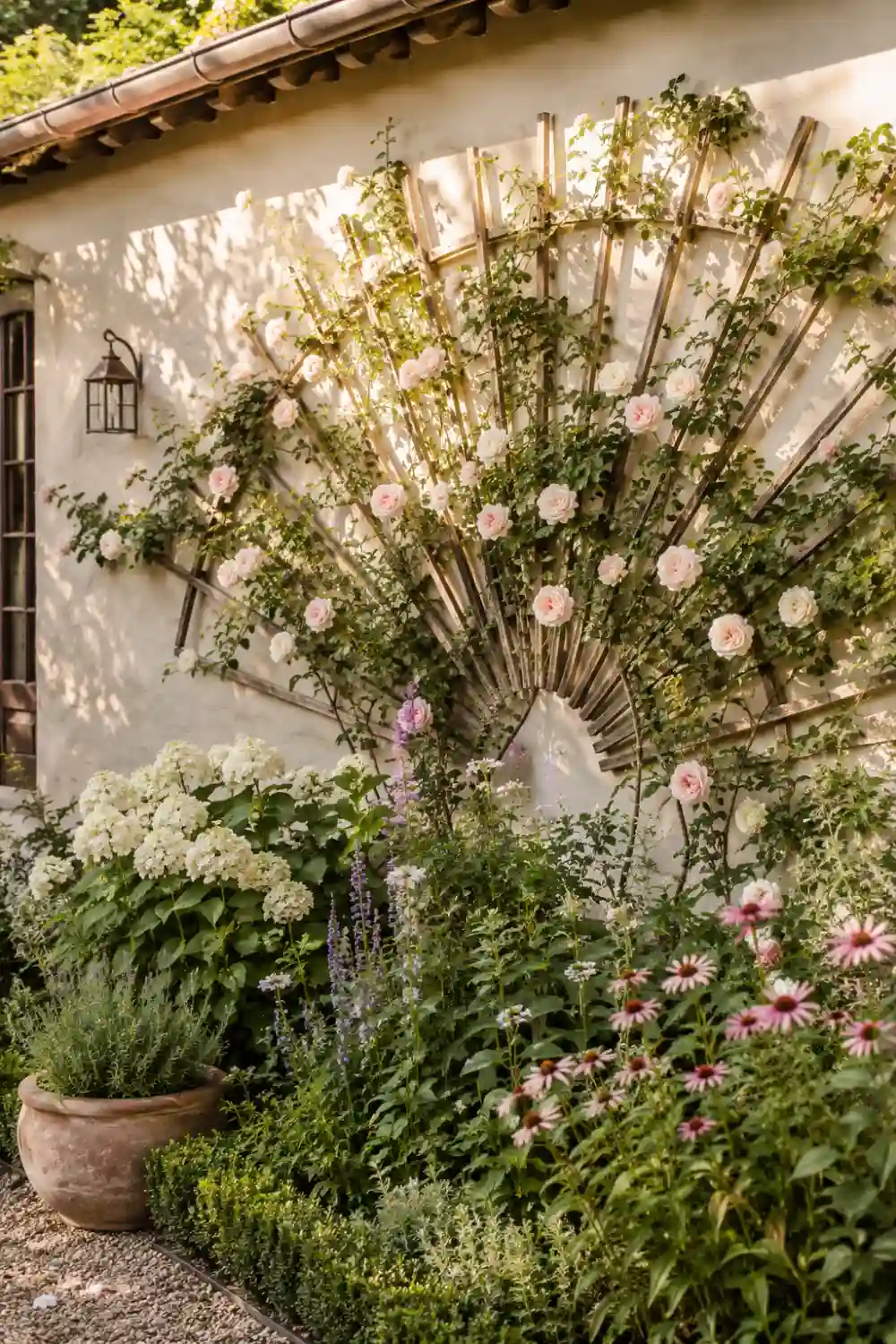 Fan-shaped wooden wall trellis covered in climbing roses and greenery mounted on a cream exterior wall above lush cottage garden flower beds in warm sunlight.