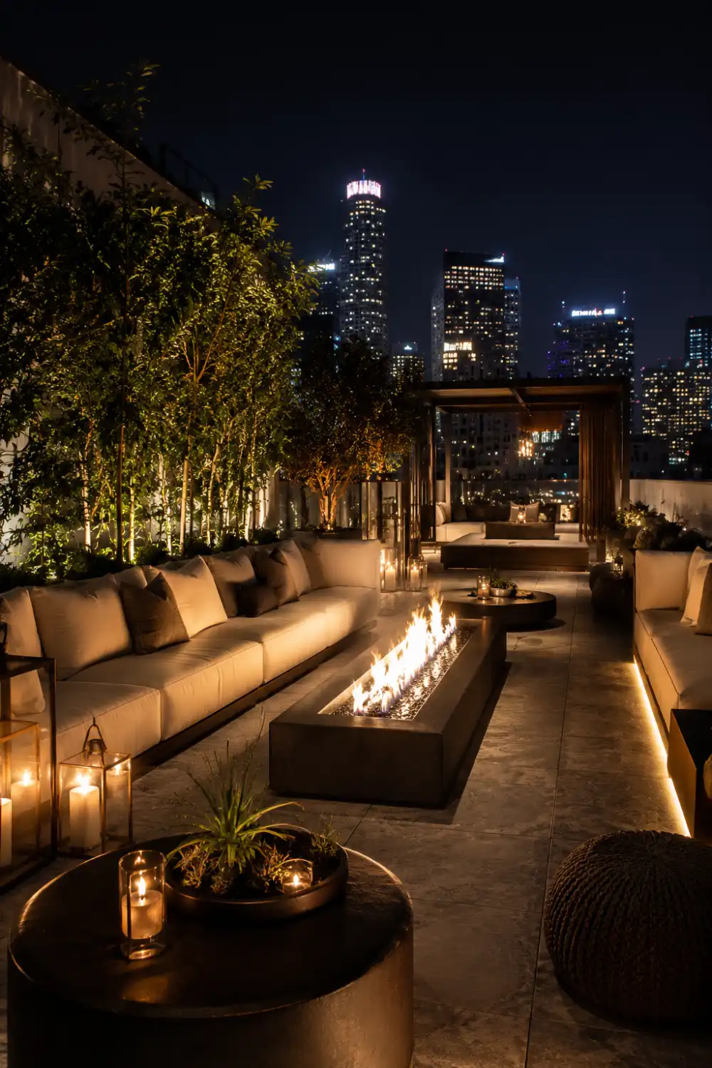 Rooftop-style fire pit lounge with linear fire table, oversized modern seating, glowing lanterns, city skyline views, warm LED lighting, and luxury outdoor patio ambiance at night.