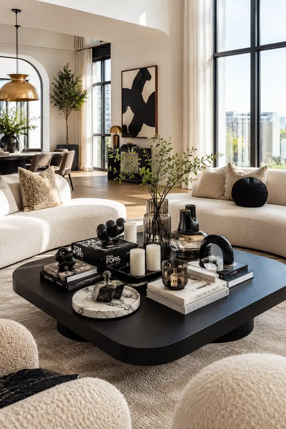 Modern luxury living room with a bold black coffee table styled using sculptural decor, designer books, candles, smoked glass accessories, and layered black-and-white accents. Cream boucle sofas, oversized windows, and soft natural lighting create a sophisticated Pinterest-worthy modern interior design aesthetic.