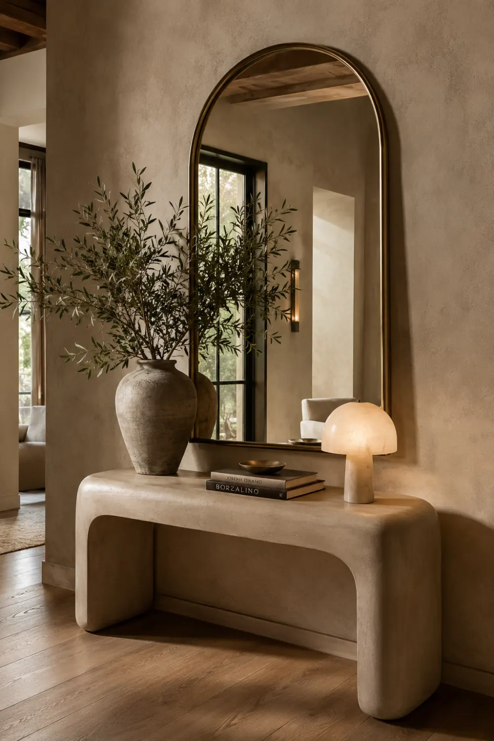 Elegant arch mirror above a curved console table with ceramic vase decor, warm glowing lamp, textured neutral walls, wood flooring, soft lighting, and luxurious modern organic entryway styling.