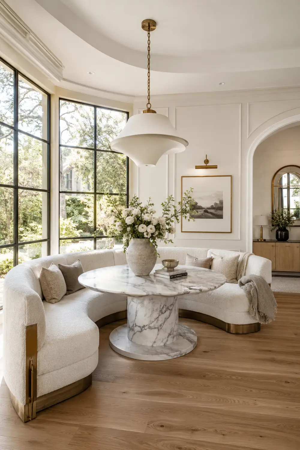 Luxury curved bouclé breakfast nook featuring a custom crescent-shaped upholstered banquette, round white marble pedestal dining table, oversized brass pendant light, floor-to-ceiling black-framed windows, fresh floral centerpiece, warm wood flooring, and elegant designer home decor in a bright modern dining corner.