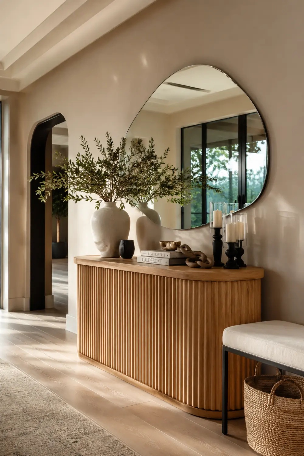 Luxury curved fluted wood console table styled with an oversized organic mirror, ceramic vases filled with olive branches, decorative candles, designer books, woven basket storage, and warm neutral decor in a bright organic modern entryway with natural wood flooring and soft sunlight.