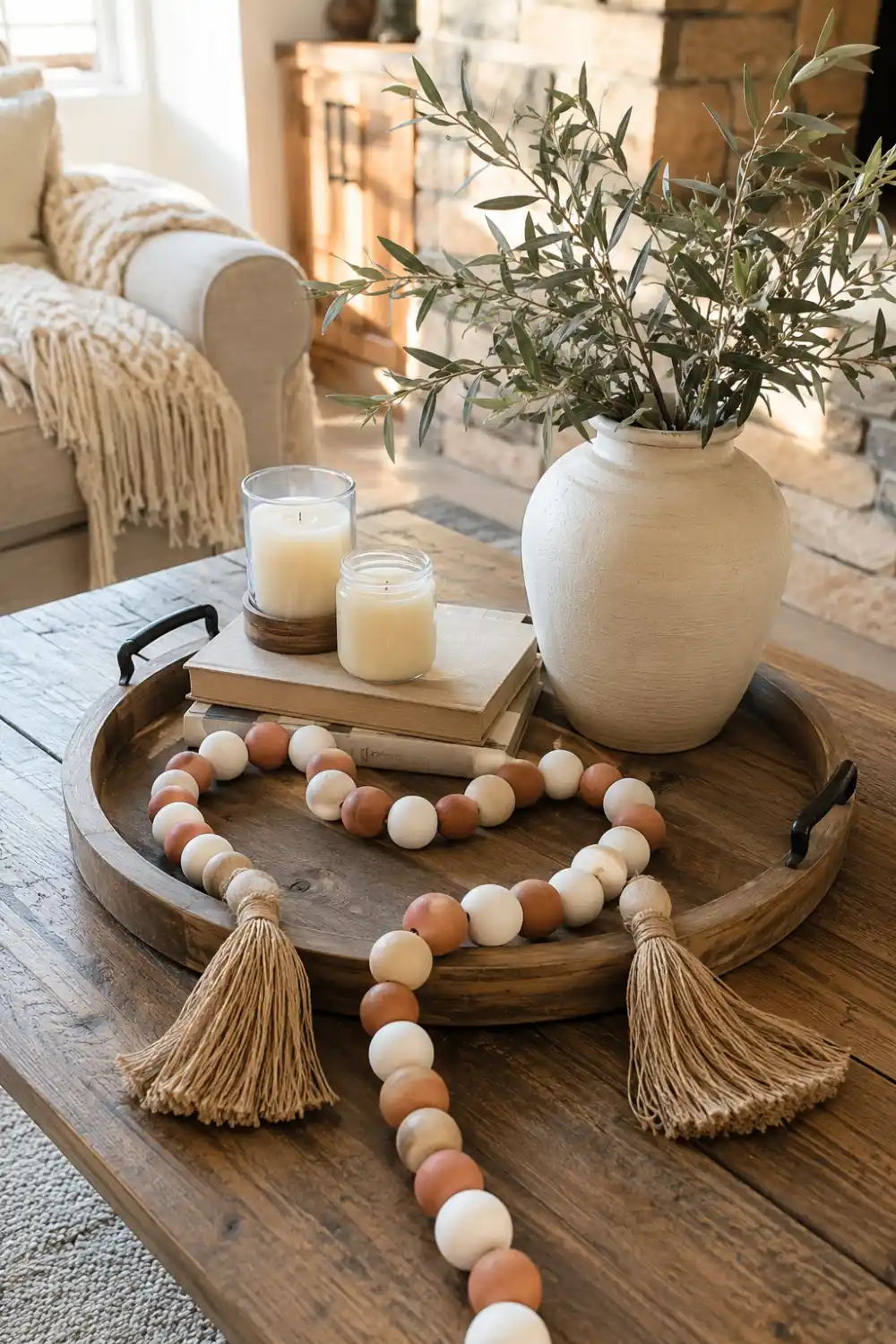 Decorative clay bead garland with white, beige, and terracotta beads and tassels draped across a wooden tray with candles, books, and a ceramic vase on a rustic coffee table.