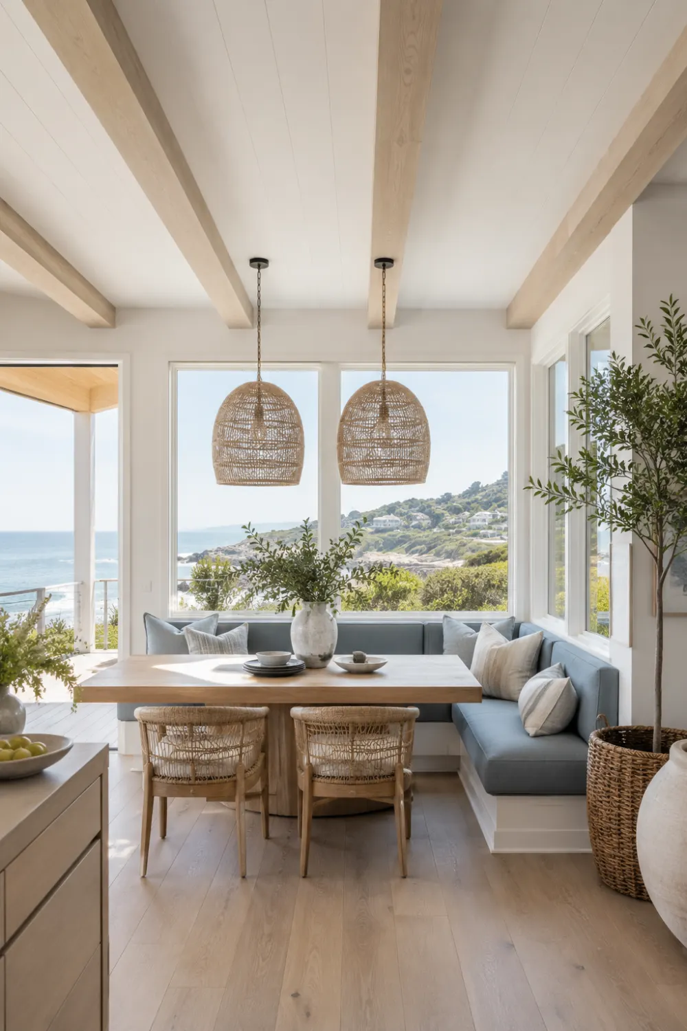 Coastal modern breakfast nook featuring soft blue built-in banquette, natural wood dining table, woven pendant lights, large ocean-view windows, wicker chairs, and bright beach-inspired home decor.