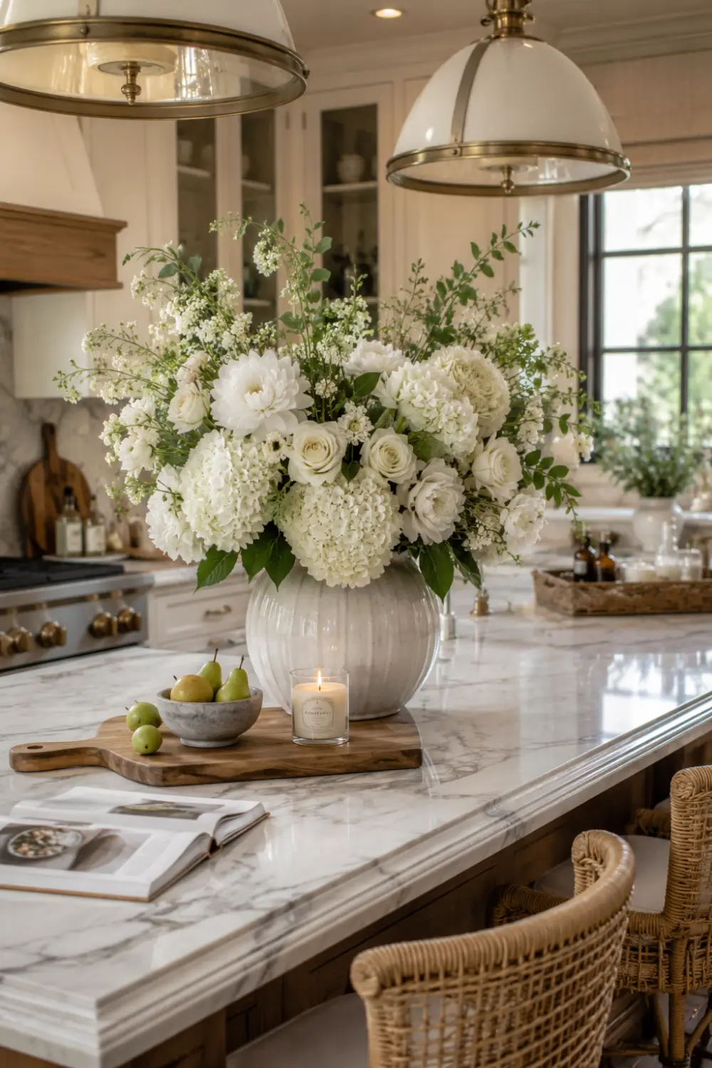 Large flower arrangement on a marble kitchen island featuring white hydrangeas, roses, greenery, candles, and decorative accents inside a bright luxury kitchen with pendant lighting.