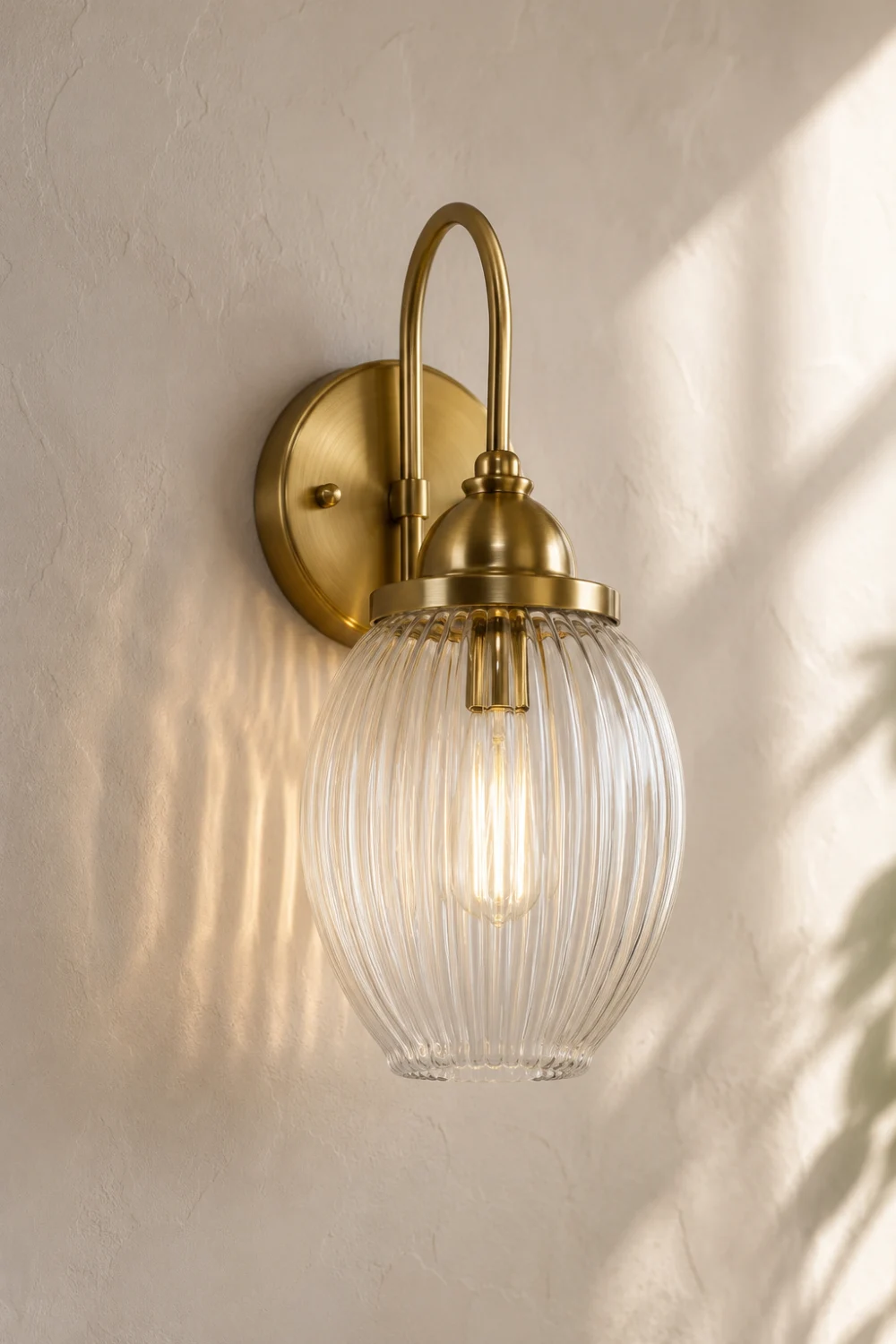 Elegant fluted glass wall sconce with a ribbed clear glass shade and brushed brass finish mounted on a textured wall, creating beautiful reflections and warm decorative lighting in a luxury interior.