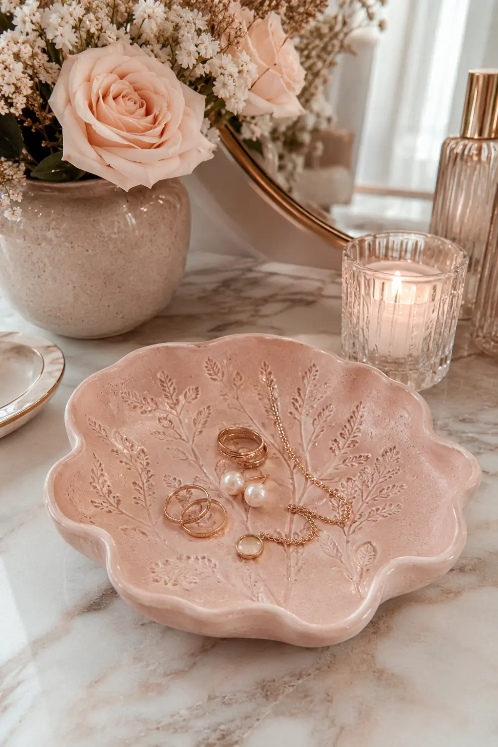 Decorative blush pink clay trinket dish with embossed botanical patterns holding rings, earrings, and jewelry on a marble vanity surrounded by candles, flowers, and luxury decorative accessories.