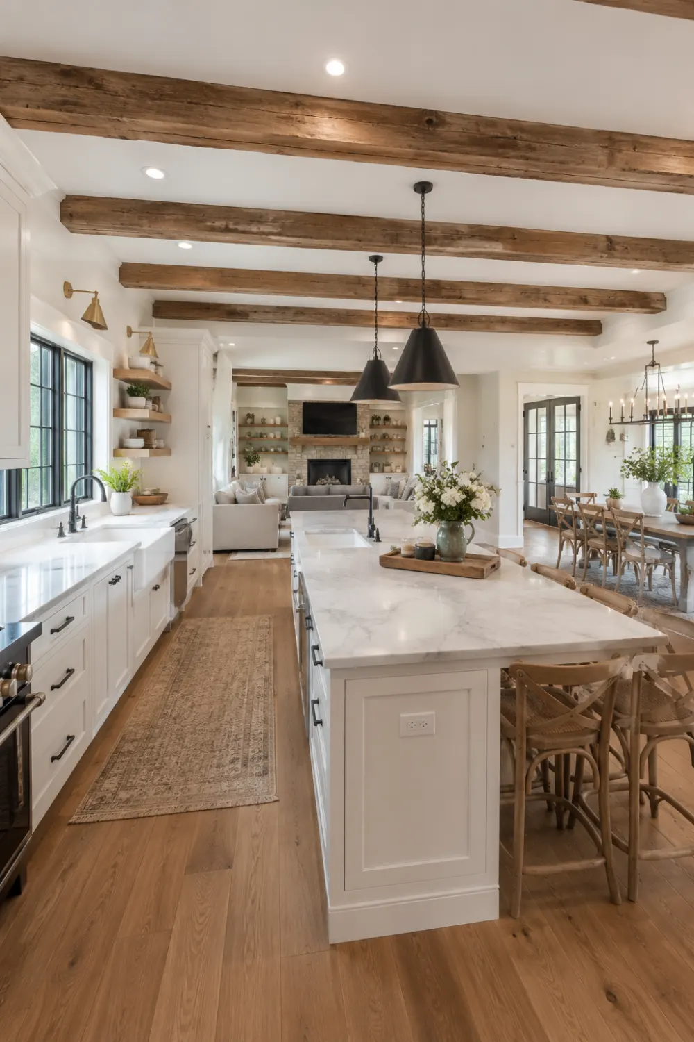 Modern farmhouse open kitchen with exposed wooden ceiling beams, large marble island, classic cabinetry, rustic wood accents, black pendant lighting, warm hardwood flooring, open shelving, and a cozy connected dining and living space.