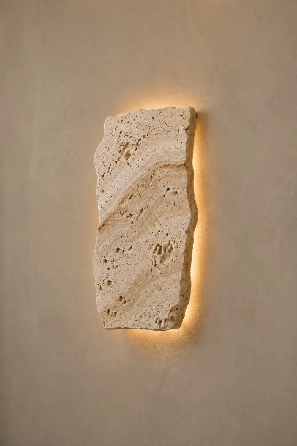 Travertine stone wall light featuring a natural textured stone slab with hidden LED backlighting, mounted on a neutral wall and creating a warm halo effect in a luxury organic modern interior.