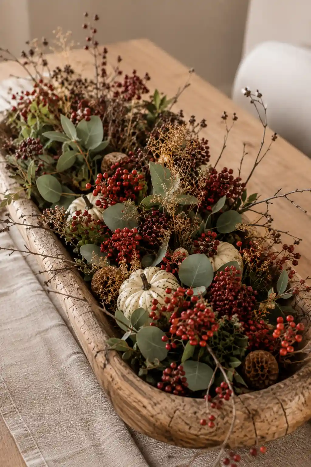 Elongated weathered wooden dough bowl filled with abundant clusters of deep red autumn berries, muted eucalyptus leaves, dried botanical stems, delicate branches, cream and speckled miniature pumpkins, natural seed pods, and layered greenery creating a rich botanical fall centerpiece on a neutral dining table.