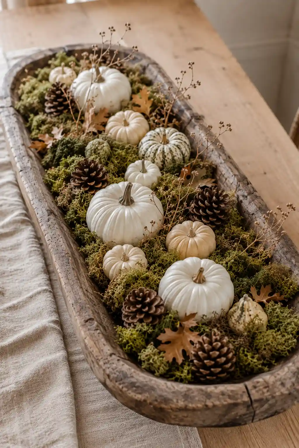 Large aged wooden dough bowl covered with lush preserved green moss and arranged with ivory, cream, and lightly speckled pumpkins, large natural pinecones, dried oak leaves, delicate branching stems, and earthy botanical accents for a soft woodland-inspired fall centerpiece on a neutral dining table.