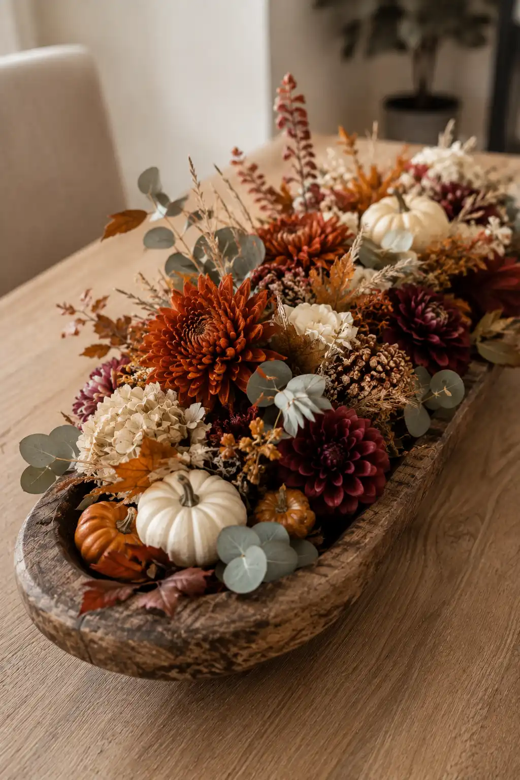 Large weathered wooden dough bowl overflowing with rich autumn flowers in burnt orange, deep burgundy, muted plum, and cream, combined with miniature white and orange pumpkins, eucalyptus leaves, dried grasses, berries, pinecones, and delicate seasonal branches on a warm wooden dining table.