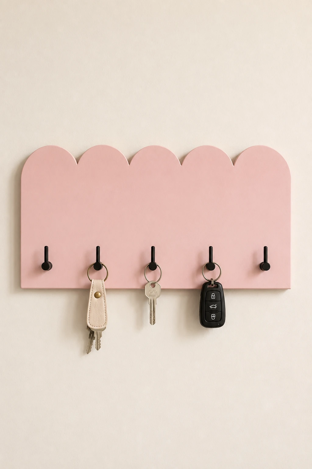 Cute scalloped DIY key holder mounted on a cream wall, featuring a soft dusty pink wooden panel, playful curved edges, five matte black hooks, leather keychain, house keys, and car keys for modern entryway organization.