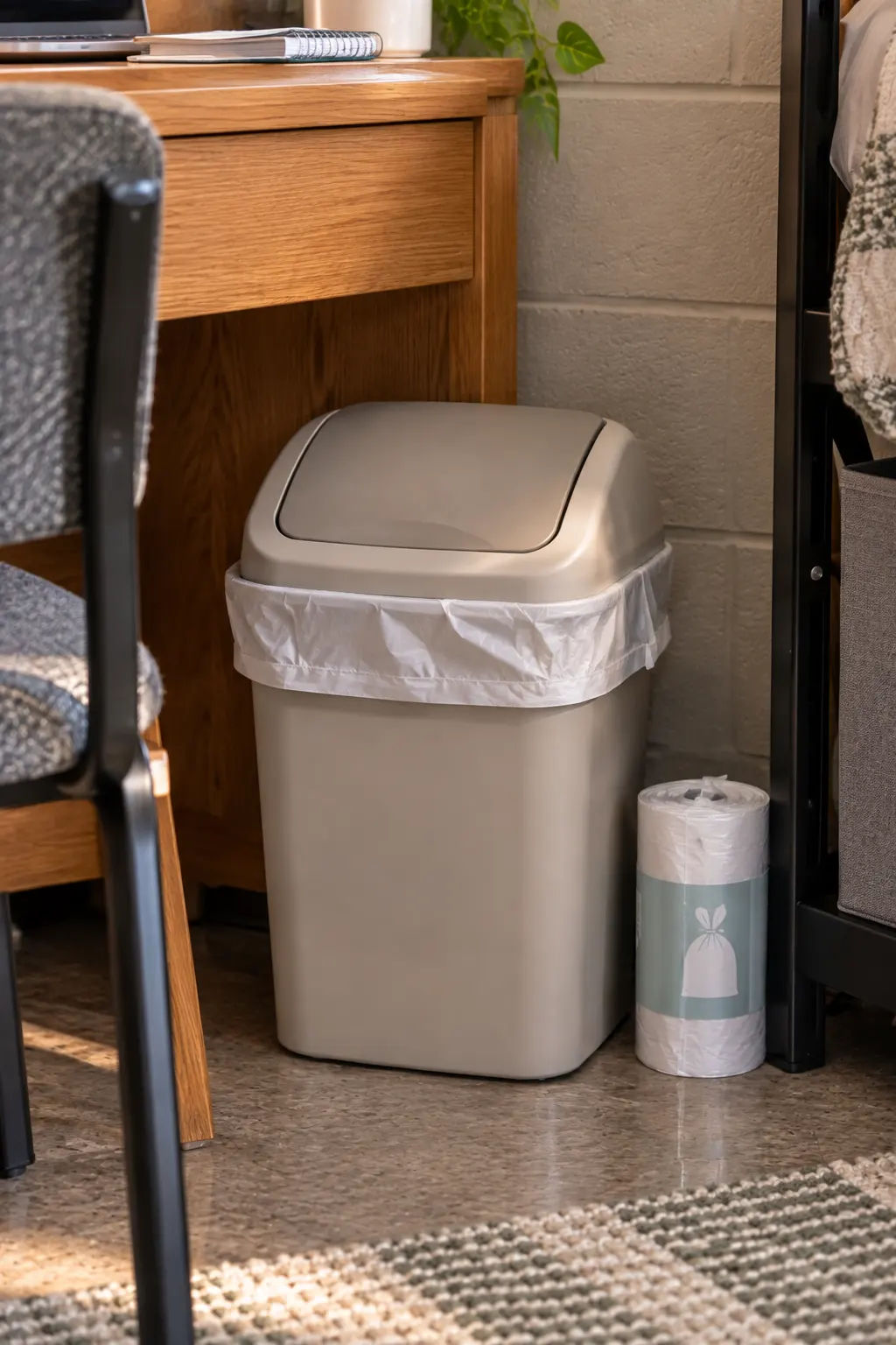 Small practical trash can with a fitted liner and replacement trash bags positioned beside a college dorm desk for convenient everyday cleanup.